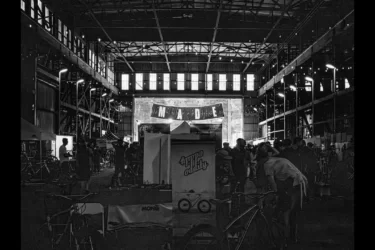 A bustling indoor event in a large, industrial space, featuring booths and displays, with people walking around and interacting. A banner displaying the word "MADE" is prominently hung at the back, illuminated by lights. Several bicycles are visible in the foreground, contributing to the vibrant atmosphere of the gathering.