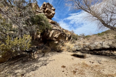 A rocky landscape featuring large boulders and dry vegetation under a partly cloudy sky. The scene includes sparse trees and low shrubs, with a sandy ground and signs of water erosion. The area conveys a rugged, natural environment. Milagrosa / Molino Basin mountain bike trail.