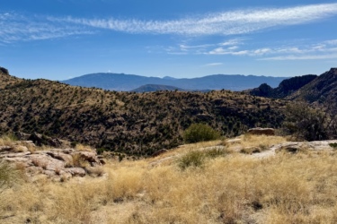 A scenic view of rolling hills and mountains under a blue sky with scattered clouds, featuring golden grasses and rocky terrain in the foreground. The landscape showcases a mix of vegetation, including shrubs and trees, set against distant mountain ranges. Milagrosa / Molino Basin mountain bike trail.