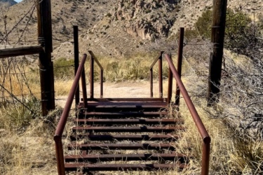 Rusty metal stairs lead down a sandy path surrounded by dry grass and shrubs, with rugged mountains in the background under a blue sky with wispy clouds. Milagrosa / Molino Basin mountain bike trail.
