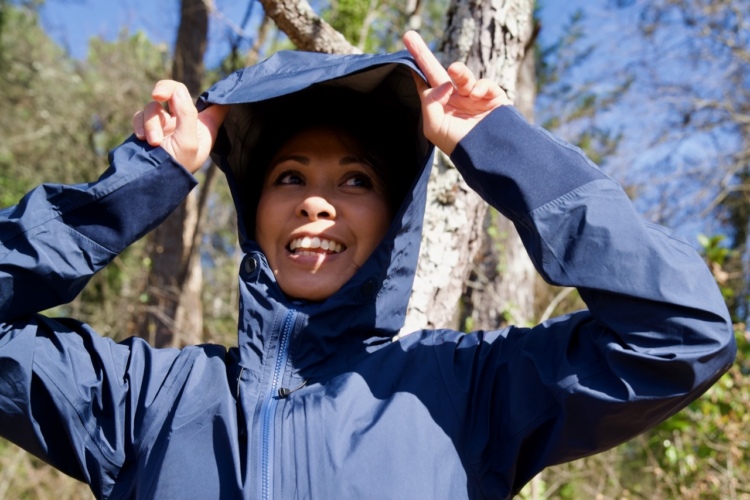 Smiling woman outdoors, adjusting the hood of her navy blue jacket, with trees and blue sky in the background.