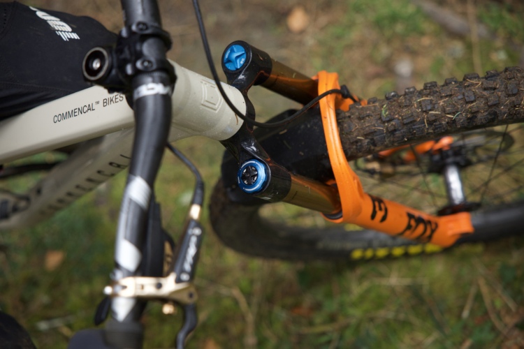 Close-up view of a mountain bike's front suspension fork and handlebars, featuring a frame labeled "Commencal Bikes." The fork has orange and black components, with blue knobs for adjustments, and the tire shows dirt, indicating off-road use. The background consists of natural greenery.