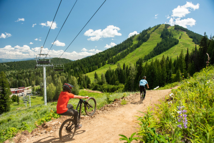 Two mountain bikers ride down a dirt trail surrounded by lush green trees and a bright blue sky with fluffy clouds. In the background, a ski lift can be seen ascending the mountain, and a lodge is visible at the base of the slope. One biker is performing a wheelie, while the other rides alongside, creating a dynamic outdoor scene.