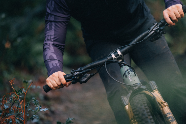 A close-up image of a cyclist's hands gripping the handlebars of a mountain bike, set against a blurred natural background. The cyclist is wearing a long-sleeved, purple jersey with a subtle brand logo on the arm. The bike features distinctive markings and is partially covered in dirt, indicating recent use on a rugged trail.