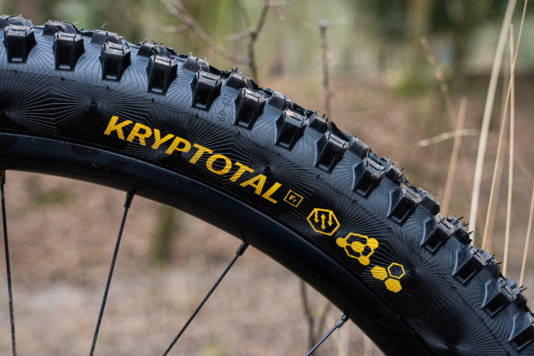 Close-up of a mountain bike tire with the text "Kryptotal" prominently displayed in yellow. The tire features a rugged tread pattern designed for off-road terrain, with visible details showing the tire's design and construction. Background includes a blurred view of natural surroundings, suggesting an outdoor setting.