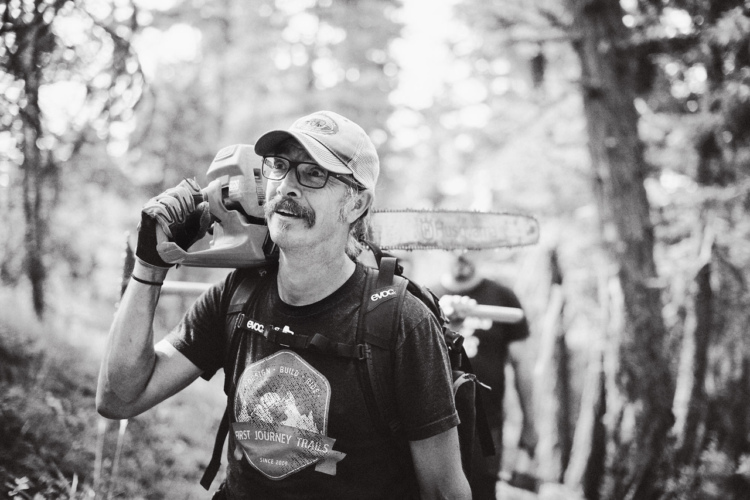 A man wearing a cap and glasses carries a chainsaw on his shoulder while walking through a wooded area. He is smiling and wearing a t-shirt with a graphic design. In the background, another person can be seen holding equipment as they work in the forest. The image is in black and white.