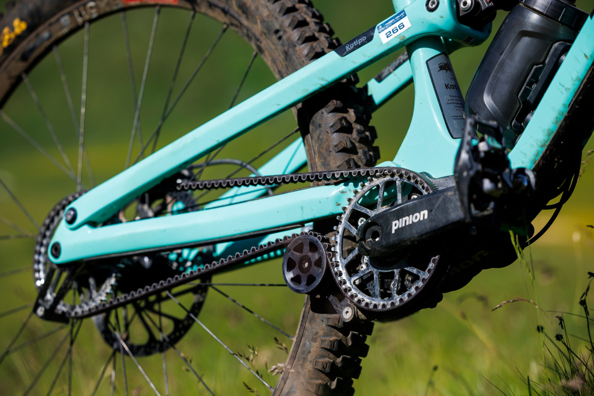 Close-up view of a turquoise mountain bike frame featuring a Pinion gearbox and chain system, set against a lush green background. The bike's rear wheel showcases rugged tires designed for off-road conditions.