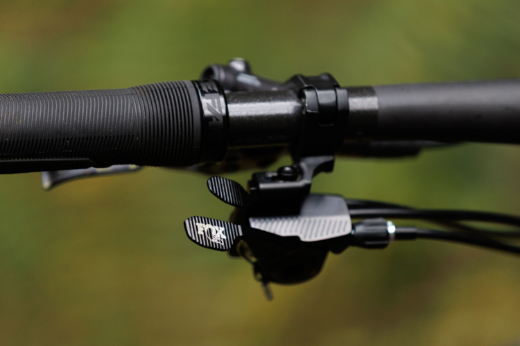 Close-up of a bicycle handlebar grip with a black lever and cable mechanism, set against a blurred green background. The handle features textured rubber for grip, and the lever showcases a distinct design with the brand logo.