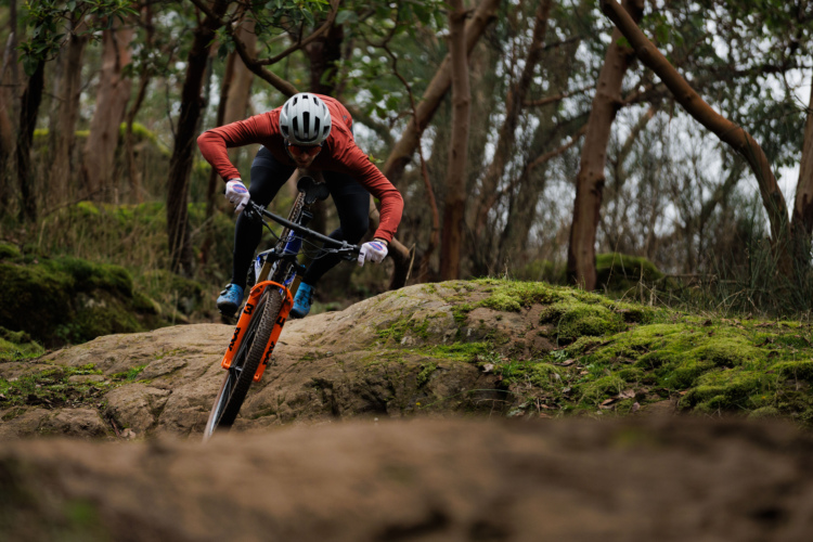 A mountain biker navigating a rocky terrain in a forested area. The rider is leaning forward on the bike, wearing a helmet and gloves, showcasing focus and determination. The ground is covered in moss and scattered rocks, with trees visible in the background.