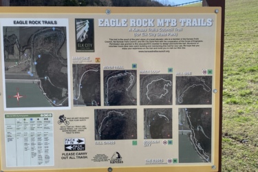 A sign displaying a map of the Eagle Rock Mountain Bike Trails located at Elk City State Park. The map features various trails, including Main Line, River Trail, Inner Loop, Hill Side, Tall Grass, Boulder City, and The Essess. It includes information about trail difficulty ratings, safety reminders, and instructions for trail users. The background shows a grassy area with trees in the distance. Eagle Rock Mountain Bike Trail mountain bike trail.