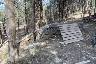 A wooden ramp is built against a large rock in a forested area, surrounded by multiple trees and underbrush. The ground is covered with leaves and small stones, and a mountain bike is parked nearby. Eagle Rock Mountain Bike Trail mountain bike trail.