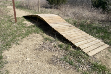 A wooden ramp made of planks, curved at the center, positioned alongside a dirt path in a natural setting with sparse grass and shrubs. Eagle Rock Mountain Bike Trail mountain bike trail.