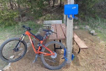 A mountain bike is parked next to a picnic table in a wooded area. A blue bike repair station is mounted on a post nearby, equipped with tools hanging from it. The ground is covered with gravel, and the surrounding trees provide a natural backdrop. Eagle Rock Mountain Bike Trail mountain bike trail.