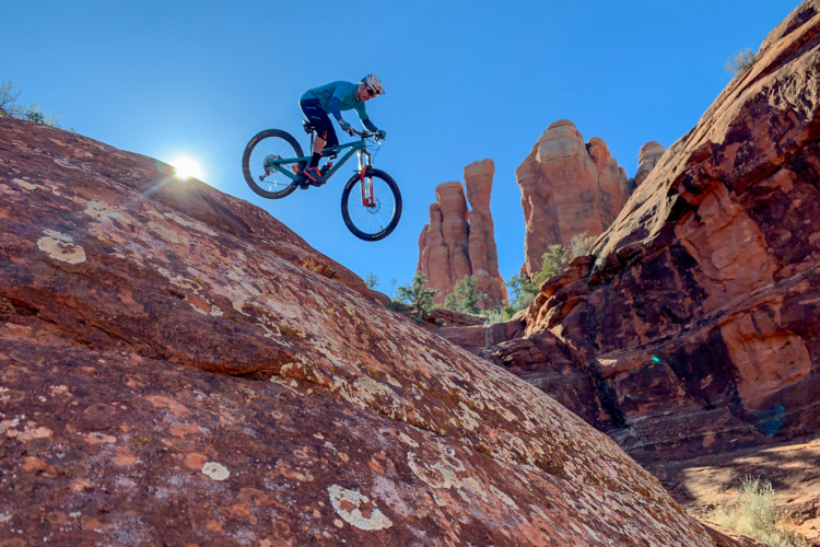 A mountain biker in mid-air, jumping off a rocky terrain with a backdrop of towering red rock formations and a clear blue sky. The sun shines brightly behind the rider, casting a dramatic light on the scene.