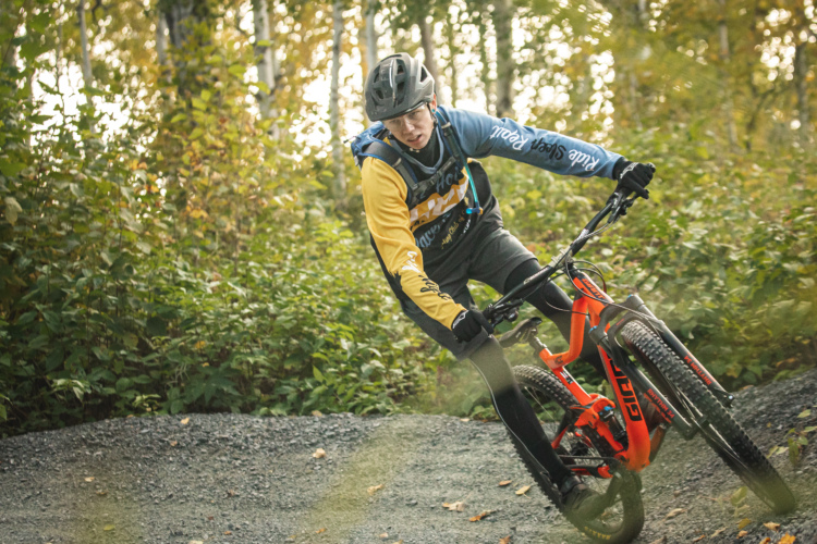 A mountain biker wearing a blue and yellow jersey and a helmet leans sharply while navigating a gravel trail surrounded by lush green foliage in an autumn setting.