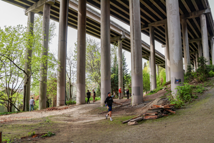 A group of people walking and socializing under a highway overpass, surrounded by trees and greenery. The scene features concrete pillars of the overpass towering above, with a mix of dirt paths and remnants of wood debris on the ground, highlighting an urban environment transitioning into nature.