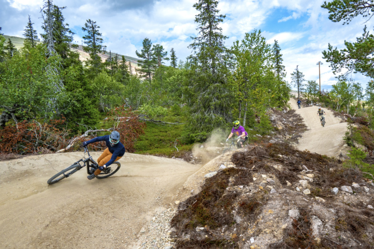 A mountain biking scene featuring three cyclists navigating dirt trails in a forested area. One rider is making a sharp turn on the left, while another is following closely behind on a separate path. The landscape includes lush green foliage and scattered rocks, with a bright blue sky overhead. Dust is being kicked up as the bikers navigate the terrain.