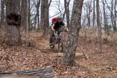 A mountain biker riding along a dirt trail in a wooded area, surrounded by bare trees and fallen leaves. The cyclist is wearing a helmet and a backpack, focusing on navigating the path while leaning forward on the bike. Bennett Gap / 138 mountain bike trail.