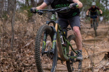 Two mountain bikers navigating a wooded trail, with one rider in the foreground riding over a log. The first rider has a beard and is wearing a helmet, a dark shirt, and shorts. The background features trees and fallen leaves, suggesting an outdoor setting in early spring. Bennett Gap / 138 mountain bike trail.