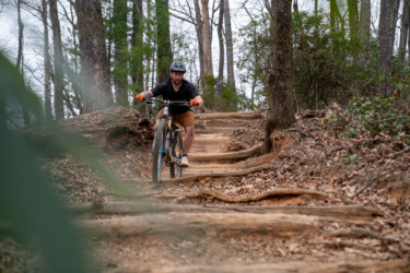 A mountain biker navigating a rocky and root-laden trail in a wooded area. The cyclist is wearing a helmet and orange gloves, focusing on the path ahead. Surrounding trees and foliage provide a natural backdrop, with fallen leaves covering the ground. Bennett Gap / 138 mountain bike trail.
