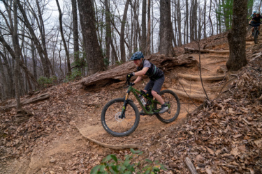 A person riding a mountain bike on a dirt trail through a wooded area. The rider is in motion, navigating a winding path with exposed roots and surrounding trees. The ground is covered in fallen leaves, and the scene appears to be set in a cool, overcast environment. Bennett Gap / 138 mountain bike trail.