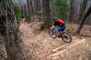 A mountain biker navigating a dirt trail surrounded by trees, with another cyclist visible in the background. The trail features loose soil, roots, and wooden features, indicating a more technical riding path. The scene captures the essence of outdoor biking adventure in a forested environment. Bennett Gap / 138 mountain bike trail.