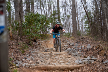 A person riding a mountain bike on a rocky trail in a wooded area, surrounded by trees and greenery. The cyclist is focused and maneuvering over the uneven terrain, with a slight incline visible in the background. Buckwheat Knob / 122 mountain bike trail.