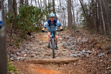A mountain biker navigating a rocky trail, with trees in the background and a log crossing the path. The rider is wearing a light blue shirt and a helmet, focused on maintaining balance while tackling the uneven terrain. Buckwheat Knob / 122 mountain bike trail.