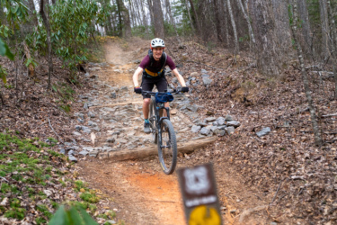 A mountain biker navigating a rocky trail in a wooded area, with visible dirt and gravel underfoot. The rider is wearing a helmet and biking gear, and there is a trail sign in the foreground indicating the path. Buckwheat Knob / 122 mountain bike trail.