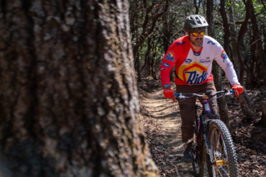 A mountain biker riding on a dirt trail surrounded by trees, wearing a colorful jersey and helmet, with sunglasses, smiling as he navigates the path. Butter Gap / Trail #123 mountain bike trail.