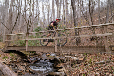 A mountain biker in a helmet rides across a wooden bridge over a stream, surrounded by bare trees and a forested landscape. Foliage is starting to bloom in the spring-like environment.