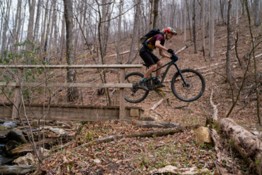A mountain biker jumps off a wooden bridge while riding through a forested area with bare trees and scattered foliage. The cyclist, wearing a helmet and a backpack, is airborne with one wheel above the bridge, showcasing an action-packed moment in nature.