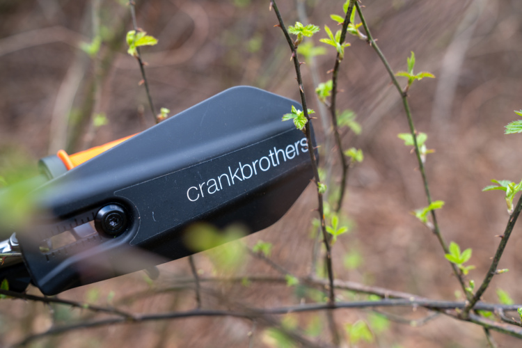 A close-up image of a tool labeled "crankbrothers" nestled among budding green leaves and thin branches in a natural setting.