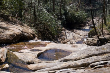 A serene stream flows over smooth, rocky terrain, surrounded by lush greenery and trees. The sunlight glimmers on the water's surface, highlighting the natural beauty of the landscape. Farlow Gap mountain bike trail.