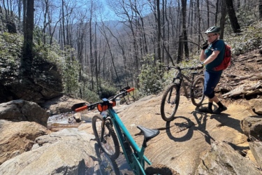 A young person wearing a helmet and a red backpack stands on a rocky path next to two mountain bikes, one green and one brown. Nearby, a small stream flows through the rocks, surrounded by trees showing early signs of spring. The background features a mountainous landscape under clear blue skies. Farlow Gap mountain bike trail.