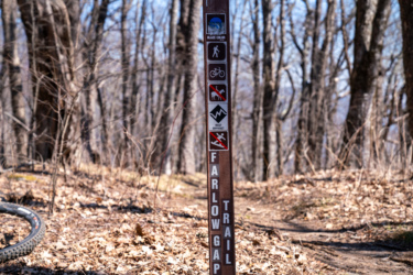 A trail signpost in a wooded area displaying various trail symbols, including indicators for hiking, mountain biking, trail difficulty, and restrictions for pets and skiing. The text "FARLOW GAP TRAIL" is visible on the sign. The ground is covered with dry leaves and the trees in the background are bare, indicating early spring. Farlow Gap mountain bike trail.