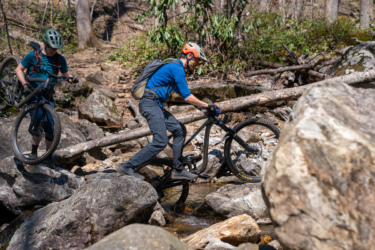 Two mountain bikers navigating a rocky stream. One rider is carefully balancing on a large rock while riding a black mountain bike, wearing a blue shirt and orange helmet. The other biker, in a green helmet and backpack, is walking beside him, carrying a bike. The scene is set in a forested area with trees and underbrush in the background. Farlow Gap mountain bike trail.