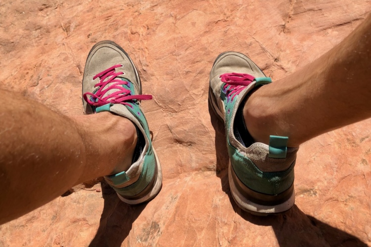 A pair of feet wearing colorful sneakers, resting on a textured reddish-brown rock surface. The shoes have turquoise and pink laces, with visible detailing on the sides and soles.
