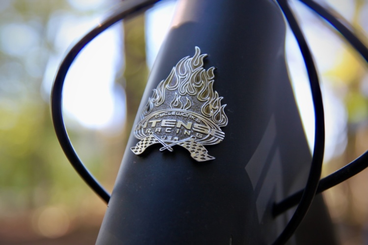 Close-up of a metallic logo featuring flames and checkered flags, attached to the frame of a bicycle. The logo reads "Cycleworks Intense Racing USA." The background is blurred with natural greenery.