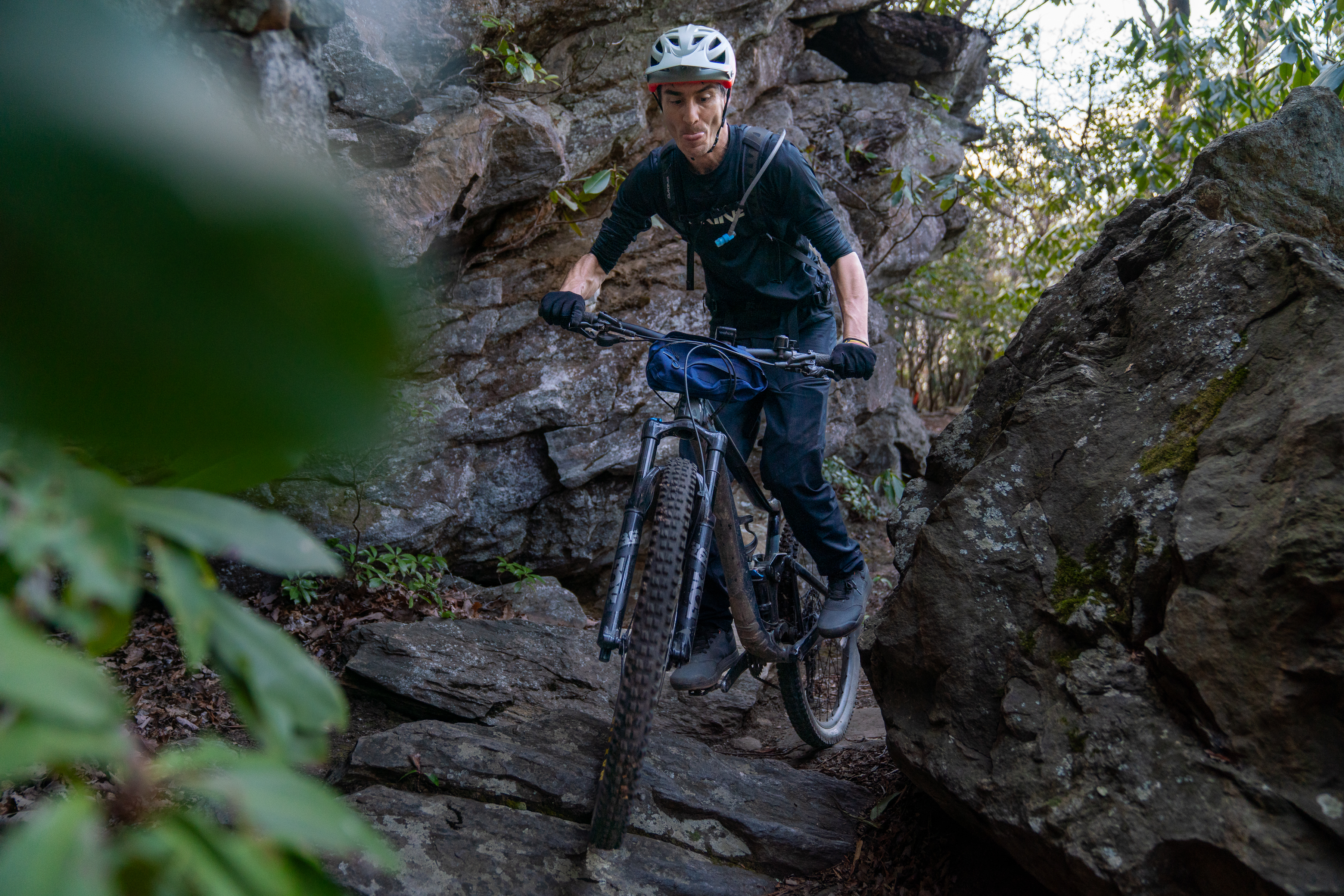 A mountain biker navigating a rocky trail surrounded by lush greenery, wearing a helmet and gloves. The rider is focusing intently as they maneuver over large stones in a wooded area. Laurel Mountain mountain bike trail.