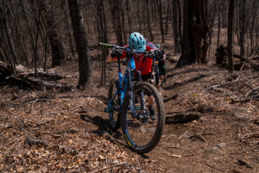 A mountain biker in a red shirt and light blue helmet is pushing their bike uphill on a rugged trail surrounded by bare trees and scattered debris. In the background, another cyclist follows closely behind. The scene captures the challenge of navigating a forested path during an outdoor biking adventure. Laurel Mountain mountain bike trail.