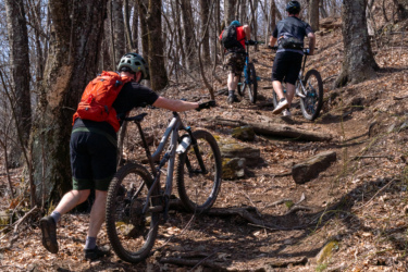 Three mountain bikers pushing their bikes uphill on a rocky dirt trail surrounded by trees, with early spring foliage on the ground. Laurel Mountain mountain bike trail.