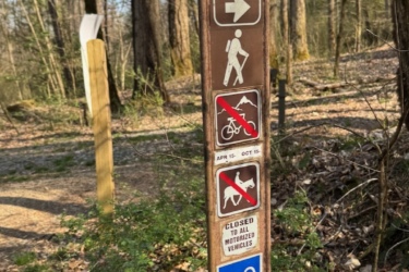 Trail sign in a wooded area with various symbols indicating trail rules and difficulty. The sign includes indicators for hiking, no bicycles, no dogs, and that the trail is closed to motorized vehicles. Additional markers show the trail’s difficulty level and direction. The background features trees and underbrush, typical of a natural trail setting. North Slope mountain bike trail.
