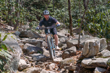 A mountain biker navigating a rocky trail in a forested area. The rider is wearing a helmet and gloves, focusing intently as they maneuver their bike over large rocks and boulders, surrounded by trees and greenery. Pilot Rock mountain bike trail.