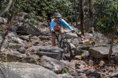 A mountain biker navigates a rocky trail in a wooded area, wearing a helmet and sunglasses. The cyclist is focused on maneuvering over large stones, with trees and greenery in the background. Another biker can be seen in the distance, also riding on the rugged terrain. Pilot Rock mountain bike trail.