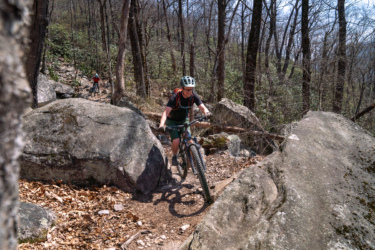 A mountain biker navigating a rocky trail in a forested area, with trees in the background and another biker visible in the distance. Pilot Rock mountain bike trail.