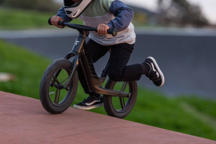 A child wearing a helmet rides a Giant Pre rCarbon balance bike on a sloped surface, focusing intently while leaning forward. The background features green grass and a blurred landscape.