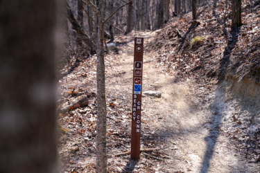 A wooden trail signpost indicating various activities, including hiking, biking, and wildlife. The sign is located along a dirt path in a wooded area, surrounded by trees and fallen leaves. The words "Searcy Conn" are visible on the sign. Searcy Creek Connector mountain bike trail.