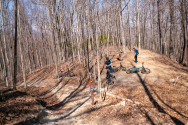 A group of four mountain bikers resting on a dirt trail in a wooded area during early spring. The trees are bare, and the ground is covered with fallen leaves. Two bikes are parked nearby, while the riders chat and relax in the sunlight. A trail marker is visible, indicating the path ahead. Searcy Creek Connector mountain bike trail.