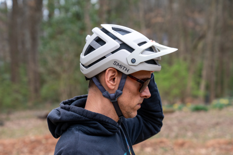 A person wearing a white bicycle helmet with ventilation features and a gray strap, standing in a wooded area. They are dressed in a dark hoodie and sunglasses, looking thoughtfully to the side.